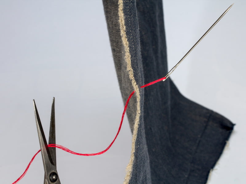 Close-up of denim being stitched with a bright red thread, needle in mid-stitch, and scissors lying nearby, symbolizing clothing manufacturing