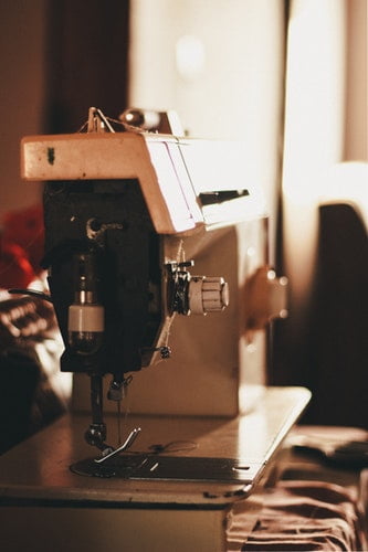 An industrial sewing machine on a workbench, illuminated by warm light, with fabrics in the background, indicative of a clothing manufacturing setting.