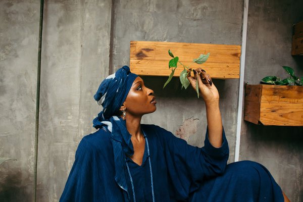 A woman in a blue traditional outfit and headwrap gently touches a green plant, symbolizing natural textiles in clothing manufacturing