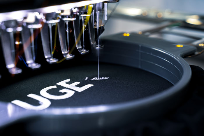 A photo of a computerized embroidery machine stitching a complex pattern onto fabric. Threads of various colors are fed into the machine.